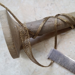 May include: A close-up of a gold mesh ribbon wrapped around a wooden dowel. The ribbon is a metallic gold color and has a woven pattern. A metal ruler is also in the image, next to the ribbon.