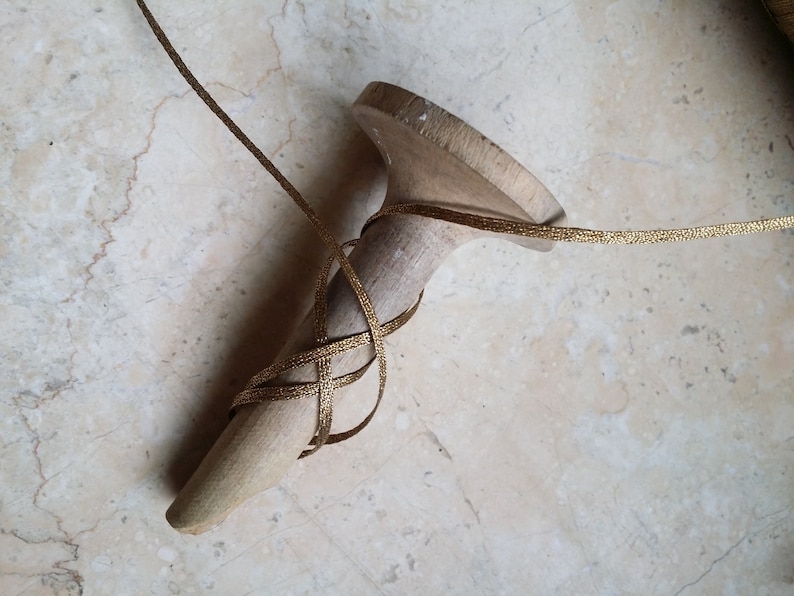 May include: A wooden shoe last with gold ribbon wrapped around it. The last is a light beige color, and the ribbon is a metallic gold. The ribbon is wrapped in a crisscross pattern around the last.