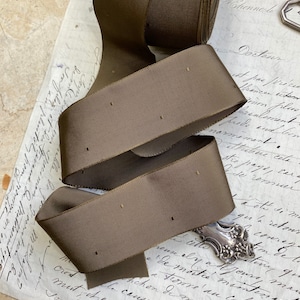May include: A roll of brown satin ribbon with a subtle dotted pattern. The ribbon is folded and arranged on a piece of paper with handwritten text.