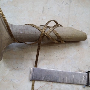 May include: A gold-colored, woven ribbon is wrapped around a wooden form. A metal ruler is positioned next to the ribbon, showing measurements in inches. The ribbon appears to be approximately 1/8 inch wide.