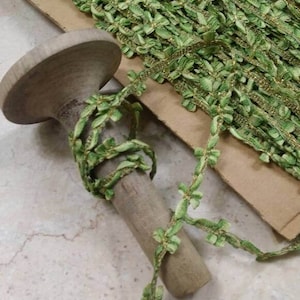 May include: Green leaf trim with gold accents, displayed on a wooden spool. The trim is a woven design with small, leaf-shaped details. A roll of the trim is visible in the background.