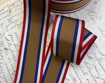 Puede incluir: Un rollo de cinta de grosgrain con una franja central ancha de color verde oliva flanqueada por rayas finas blancas, azules y rojas en ambos bordes. La cinta tiene una superficie texturizada y acanalada, presentada en forma de bobina con un extremo suelto.