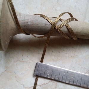May include: Close-up of a gold metallic ribbon wrapped around a wooden dowel. A metal ruler is placed next to the ribbon. The ribbon has a woven texture and is a warm gold color. The dowel is light brown.