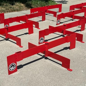 Könnte beinhalten: Eine Sammlung leuchtend roter Metallhürden, die für das sportliche Training bestimmt sind. Jede Hürde hat einen rechteckigen Rahmen mit einem markanten Logo an der Seite. Die Hürden sind auf einer Betonoberfläche angeordnet, was auf eine Verwendung im Freien hindeutet.