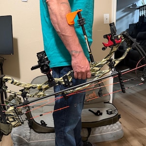 May include: A compound bow with a camouflage pattern, featuring a black sight, a red arrow rest, and bright orange cables. The bow includes a black stabilizer rod labelled Stinger with an orange end piece, held horizontally by a hand.