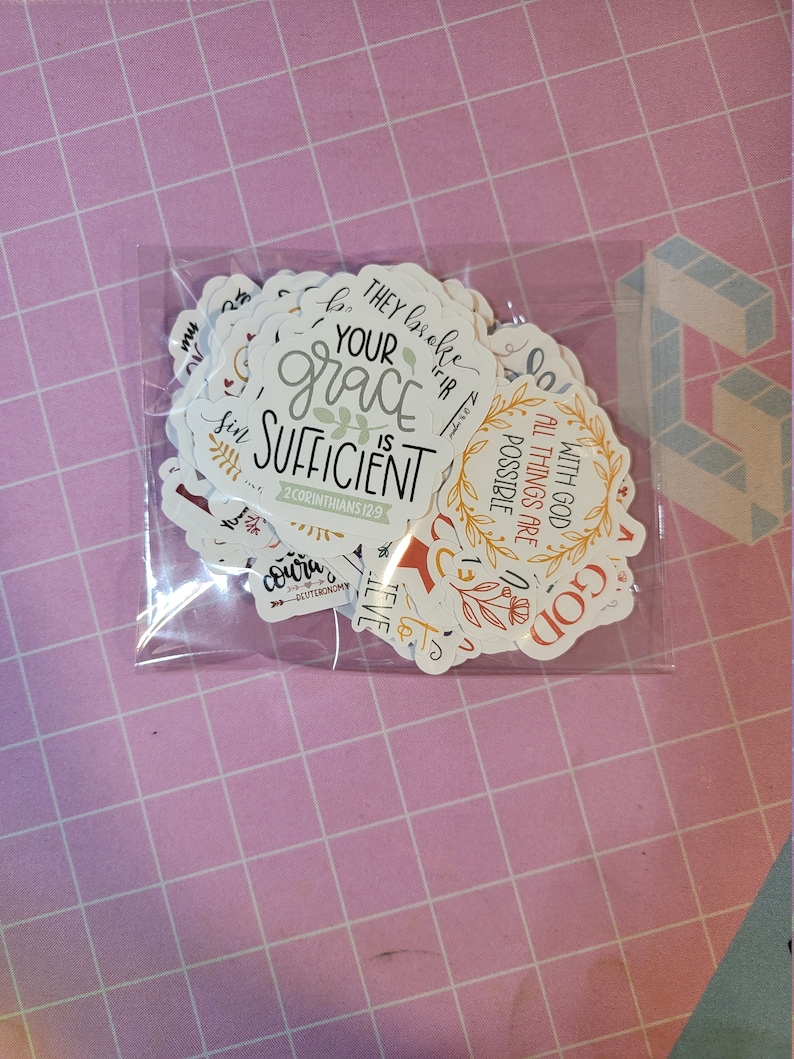 May include: A clear plastic bag containing a collection of white stickers with various inspirational quotes and floral designs. The stickers feature phrases like "Your grace is sufficient" and "With God all things are possible."