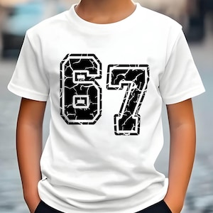 May include: White t-shirt with the number "67" in a distressed black font. The shirt is worn by a young person with blond hair, standing outdoors. The background is a blurred street scene.