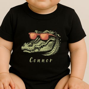 Op de afbeelding: Zwarte babybody met een groen-gele alligator met een rode zonnebril. De naam "Connor" is in lichtgroen schrift onder de alligator-afbeelding gedrukt. De body heeft korte mouwen.