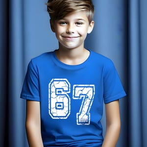 May include: A royal blue t-shirt with the number "67" in white, featuring a cracked design. The shirt is short-sleeved and worn by a person with light brown hair, smiling in front of a blue backdrop.