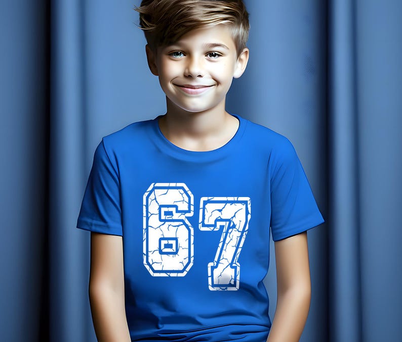 May include: A royal blue t-shirt with the number "67" in white, featuring a cracked design. The shirt is short-sleeved and worn by a person with light brown hair, smiling in front of a blue backdrop.