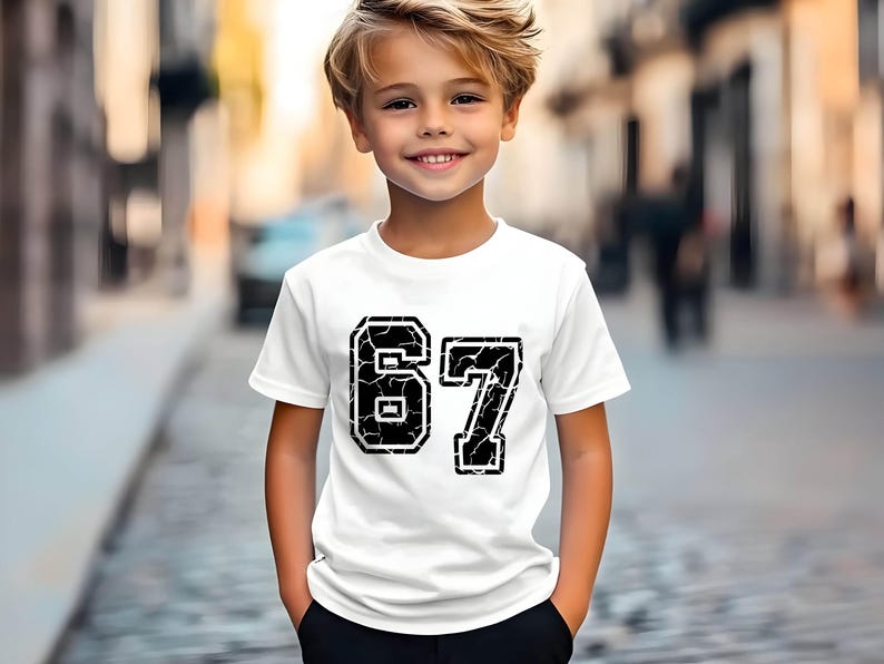 May include: White t-shirt with the number "67" in a distressed black font. The shirt is worn by a young person with blond hair, standing outdoors. The background is a blurred street scene.