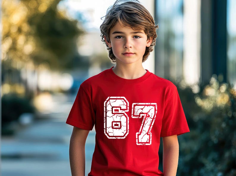 May include: A red t-shirt featuring the number "67" in a distressed, cracked white font. The short-sleeved shirt is worn by a person with light brown hair, set against a blurred outdoor background. The shirt is a casual apparel item.