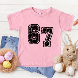 May include: Pink toddler t-shirt with the number 67 in a distressed black font. The shirt is displayed with a denim garment, a nest with Easter eggs, a stuffed bunny, and carrots.