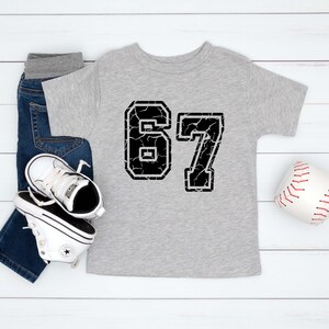 May include: A light gray toddler t-shirt with the number 67 in a distressed black font. The shirt is paired with blue jeans, white sneakers, and a baseball. The items are arranged on a white wooden surface.