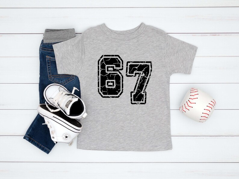 May include: A light gray toddler t-shirt with the number 67 in a distressed black font. The shirt is paired with blue jeans, white sneakers, and a baseball. The items are arranged on a white wooden surface.