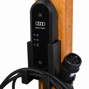 Op de afbeelding: Een zwarte Audi e-tron oplaadeenheid voor elektrische voertuigen, gemonteerd op een houten paal. Het apparaat heeft een verticaal bedieningspaneel met het Audi-logo, e-tron tekst en statuspictogrammen. Een dikke zwarte kabel is opgerold.