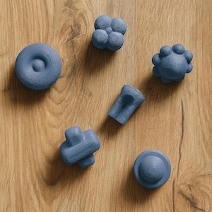 May include: Six blue, abstract, 3D-printed objects are arranged on a light brown wooden surface. The objects have different shapes, including a disc, a cube, a cylinder, and a cross-like form. The shapes are all in a blue hue.
