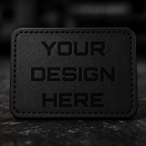 May include: A black rectangular patch with the words "YOUR DESIGN HERE" embossed on the surface. The patch has rounded corners and a stitched border. The background is a blurred, dark surface.