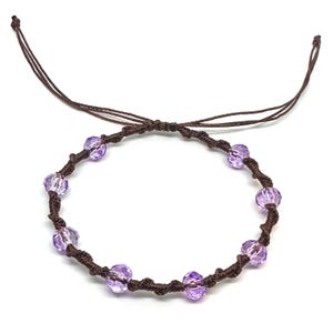 May include: Adjustable bracelet featuring faceted lavender beads strung on a dark brown, knotted cord. The bracelet has a sliding knot closure for a custom fit. The beads are evenly spaced around the cord.
