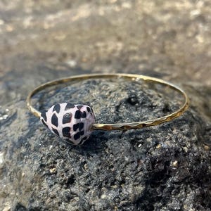 May include: A gold-colored bangle bracelet featuring a natural seashell with black and white markings. The bracelet has a hammered texture and is resting on a dark, textured rock surface. The seashell is the focal point of the jewelry.