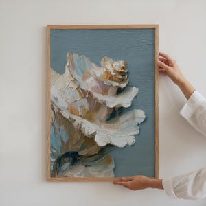 May include: A framed floral painting with a light wooden frame. The artwork features a large, textured flower in shades of white, cream, and peach against a muted blue-grey background. The painting is held by a person wearing a white shirt.