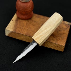 May include: A small, handcrafted knife with a light wood handle and a silver blade. The knife rests on a wooden block with a decorative wooden object in the background. The blade is sharp and pointed, suggesting a tool for detailed work.