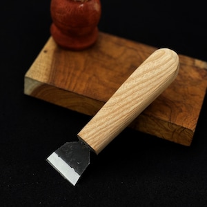 Könnte beinhalten: Ein Holzbeitel mit hellem Holzgriff und silberner Klinge. Der Beitel liegt auf einem kleinen Holzblock, mit einem kleinen, roten, abgerundeten Objekt im Hintergrund. Der Hintergrund ist schwarz.