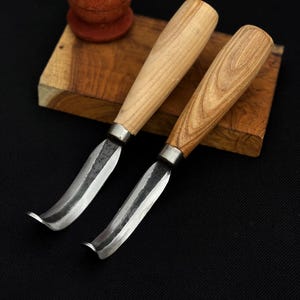 May include: Two wood carving knives with curved blades and wooden handles. The blades are silver and the handles are light brown. A small, round, reddish-brown object sits on a wooden block in the background.