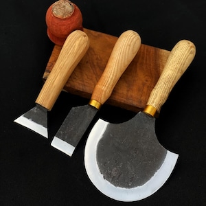 May include: Three leatherworking tools with wooden handles and metal blades. The blades have different shapes: a triangle, a rectangle, and a half-moon. The handles are light-colored wood, and the blades are dark metal.
