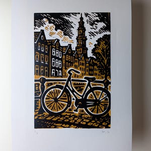 May include: Black and gold linocut print of a cityscape with a bicycle in the foreground. The print features buildings, a church tower, and a tree. The bicycle is black with gold wheels. The sky is black and white.
