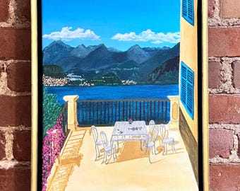 May include: A framed painting depicts a scenic outdoor terrace overlooking a lake and mountains under a bright blue sky. The terrace features a white table and chairs, with pink flowers and a railing. Blue shutters are visible on the side of the building.