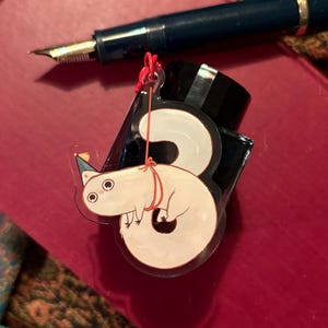 Può includere: Un ciondolo bianco a forma di numero tre, in stile cartone animato, con un cappello da festa e un filo rosso, accanto a un flacone di inchiostro nero e una penna stilografica blu. Il ciondolo è su una superficie rossa.