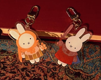 May include: Two acrylic keychains featuring cartoon bunnies. One bunny wears an orange outfit and holds flowers, the other wears a red outfit and holds a sword. Both have gold-colored clasps.