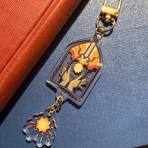 May include: A clear acrylic keyring with a gold-coloured clasp. The keyring features a stained-glass design with hands reaching for a sunburst. A smaller sunburst charm hangs below. The background is a blue surface.