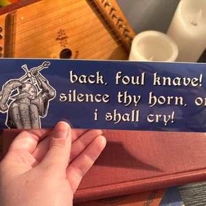 Może przedstawiać: Niebieska prostokątna naklejka z białym tekstem: „back, foul knave! silence thy horn, or i shall cry!”. Czarno-biała ilustracja rycerza z mieczami po lewej stronie. Naklejka jest na czerwonej książce.