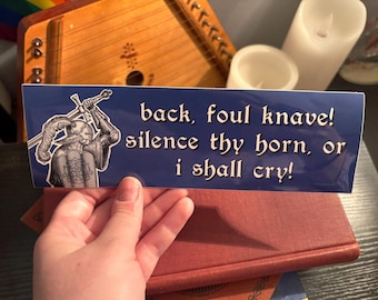 Peut inclure: Autocollant rectangulaire bleu avec texte blanc : « back, foul knave! silence thy horn, or i shall cry! » Illustration en noir et blanc d'un chevalier avec des épées à gauche. L'autocollant est sur un livre rouge.