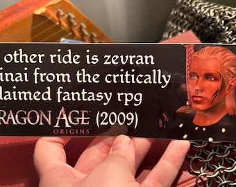 May include: A black bumper sticker with white text that reads "my other ride is zevran arainai from the critically acclaimed fantasy rpg Dragon Age (2009) Origins." Features a character portrait. The sticker is held by a hand.