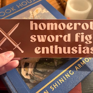 May include: A brown rectangular sticker with the words "homoerotic sword fight enthusiast" in a light brown font. Two crossed swords are depicted in the upper left corner. The sticker is held over a blue book.