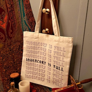 Puede incluir: Un bolso tote color crema con el texto "INVENTORY IS FULL" impreso en un patrón repetido. La bolsa cuelga de una pared de madera con ganchos de latón. Una espada decorativa y libros están en primer plano.