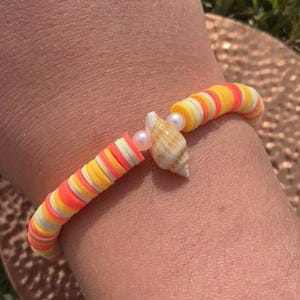 Peut inclure: Un bracelet coloré composé de perles en forme de disque orange, jaune et blanc. Le bracelet est orné d'un petit coquillage et de deux perles. Le bracelet est porté au poignet.