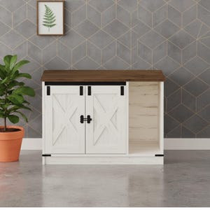 May include: A white cabinet with a dark brown top and a sliding door design. The cabinet has two doors with an X-shaped design and black hardware. A framed fern print hangs on the wall above the cabinet. A potted plant sits to the left.