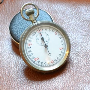 Könnte beinhalten: Eine Vintage-Stoppuhr mit weißem Zifferblatt, schwarzen Zeigern und einem goldfarbenen Metallgehäuse. Die Zahlen sind schwarz und rot. Die Stoppuhr liegt auf einer braunen Lederoberfläche, mit einem schwarzen Lederetui im Hintergrund.
