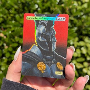 May include: A rectangular trading card featuring a holographic top banner with the text Caesar, Legion's Emperor and mana symbols. The illustration depicts a figure in a dark metallic helmet and armour against a vibrant red background.