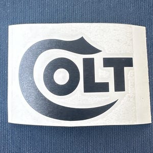 May include: A white sticker with the word "COLT" in black, featuring a stylized "C" design. The sticker is rectangular and has a slightly textured surface. The background is a solid blue fabric.