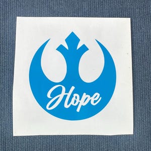 May include: A white square sticker featuring a blue emblem with the word "Hope" in a cursive font. The emblem is a stylized design with a central diamond shape and curved elements, set against a blue background.