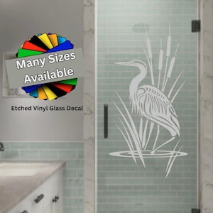 May include: An etched vinyl glass decal featuring a white graphic of a heron standing among tall reeds and cattails. The decal is applied to a glass shower door. A colorful circular graphic with the text Many Sizes Available is present.
