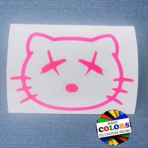 May include: A bright pink vinyl decal featuring a stylised cat face outline with X-shaped eyes and three whiskers on each side. A circular graphic in the bottom right corner displays a rainbow fan of colours with the text MANY COLORS TO CHOOSE FROM.