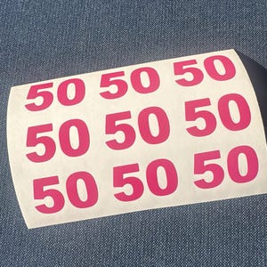 May include: Sheet of stickers with the number "50" printed in bright pink. The stickers are arranged in three rows of three, with the number repeated nine times. The stickers are on a white background.