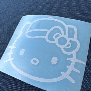May include: A white vinyl decal featuring the outline of the character Hello Kitty wearing a baseball cap. The design includes the iconic bow on the side of the cap, two oval eyes, a small oval nose, and three whiskers on each side of the face.
