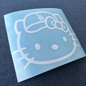 May include: A white vinyl decal featuring the outline of the Hello Kitty character wearing a baseball cap with the letters LA printed on the front. The design includes the character's signature bow on the side and whiskers on both cheeks.